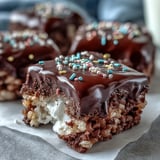 Freshly cut squares of Chocolate Covered Rice Krispy Treats stacked on a white plate, a perfect sweet snack for bake sales or a family movie night.